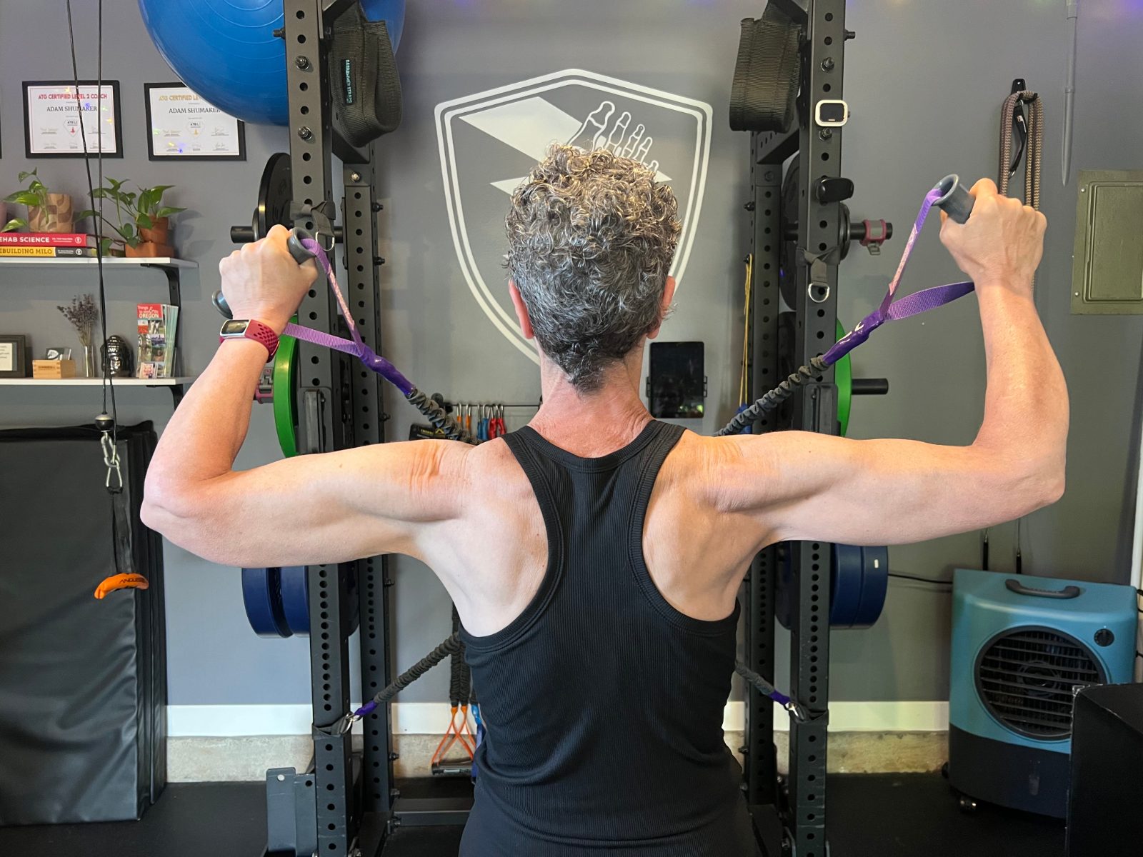 A client working through a resistance-band pulling exercise in the No Easy Feet gym, facing the studio's Foot-Shield logo on the wall.